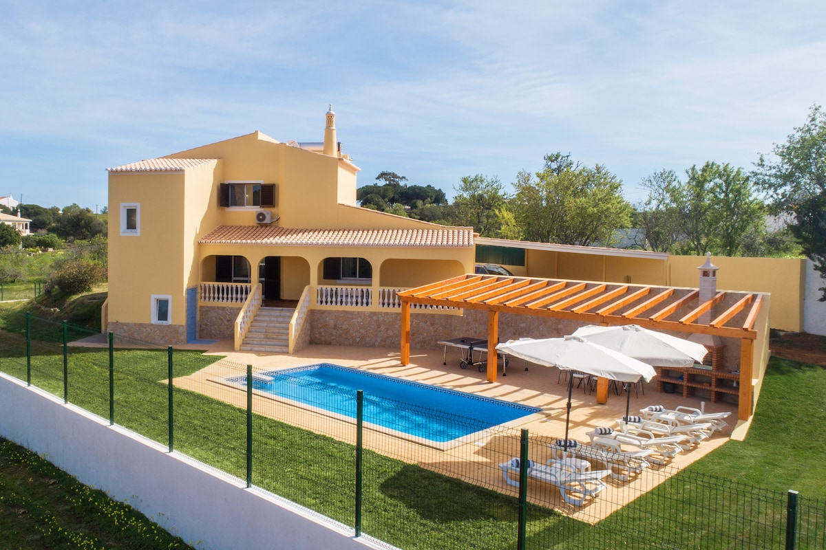 An overview of the villa reveals a two-story yellow structure with a spacious private saltwater pool and sun loungers. A shaded area with a wooden pergola provides seating for outdoor dining. Lush greenery surrounds the property, enhancing the outdoor space.
