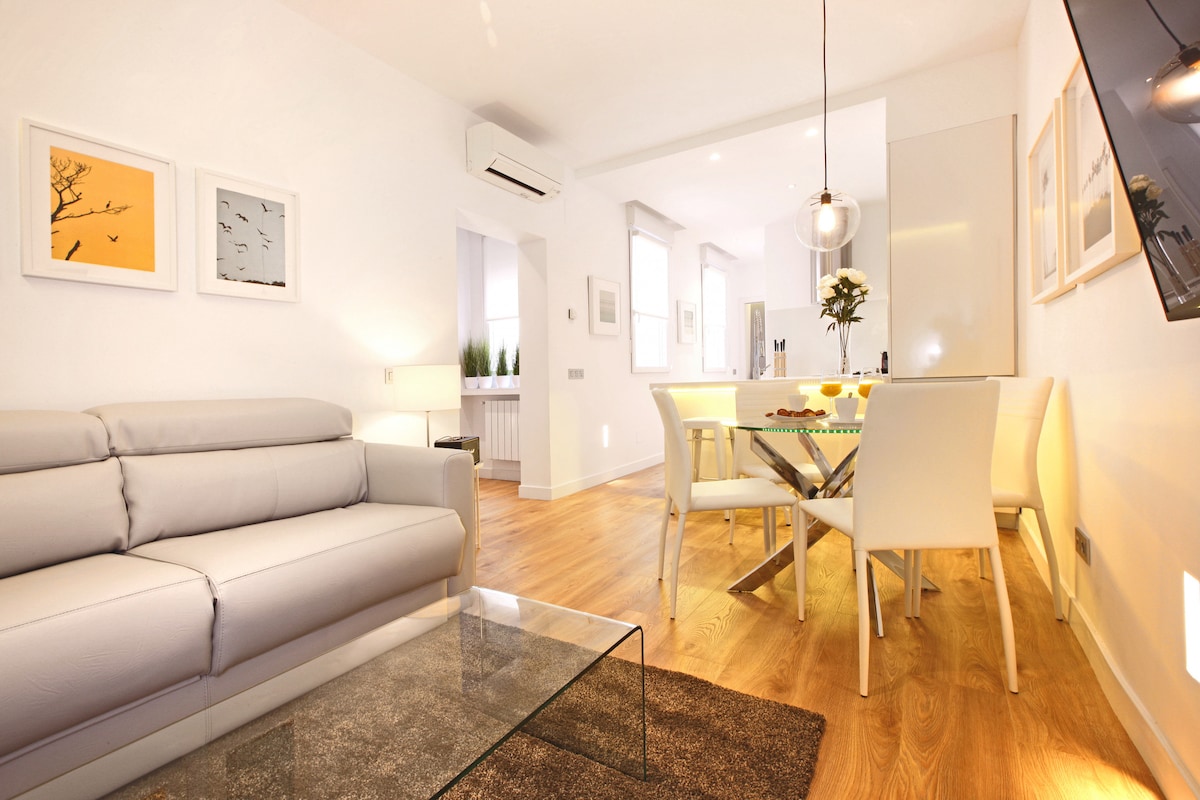 A modern living area features a light-colored sofa and a glass coffee table placed on a soft rug. A dining table with white chairs is positioned beside a well-equipped kitchen, illuminated by natural light from nearby windows.