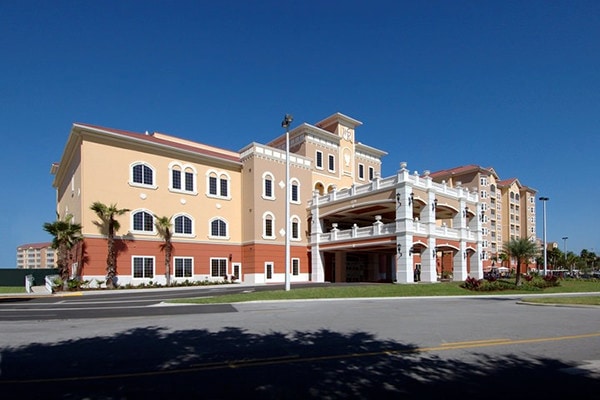 The exterior of a multi-story building features a blend of light yellow and orange façades, complemented by white architectural elements. Palms and manicured shrubs enhance the landscape. A covered entryway invites guests into the lobby, set under a clear blue sky.