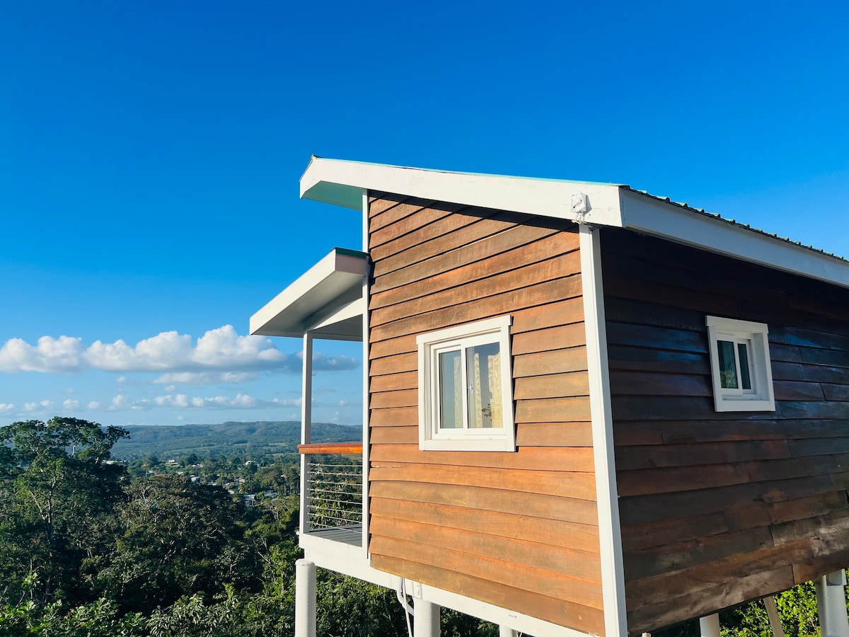 The exterior of the guesthouse is shown, featuring wooden paneling and a slanted roof. A small balcony protrudes from the structure, overlooking a lush landscape under a clear blue sky, enhancing the feeling of tranquility.