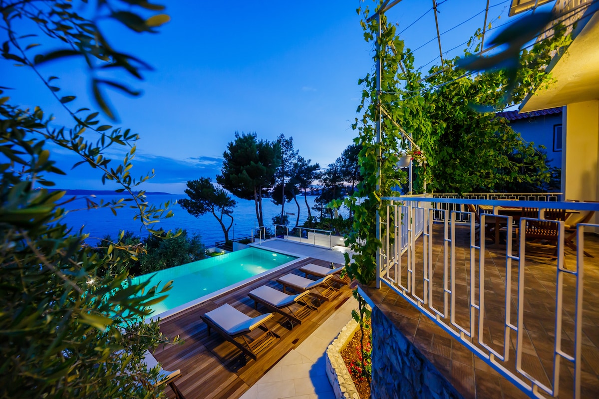 A view of the infinity swimming pool, which features wooden decking and sun loungers, is framed by greenery. The tranquil sea is visible in the distance under a soft evening sky, and a cozy terrace area can be seen, complementing the inviting outdoor space.
