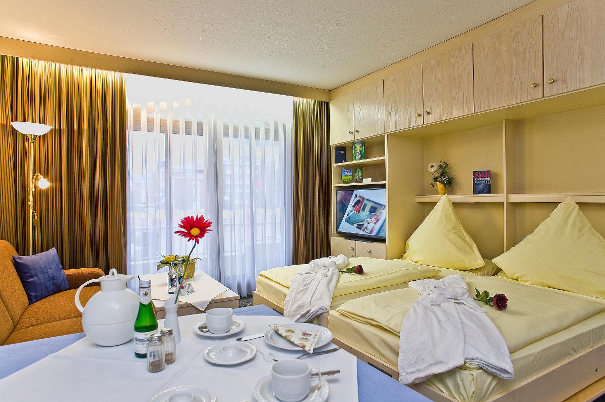 The image displays a cozy room featuring twin beds with yellow blankets and decorative pillows. A comfortable seating area is located beside a window adorned with sheer curtains. A table is set with a tea service and two glasses of water, adding a welcoming touch.