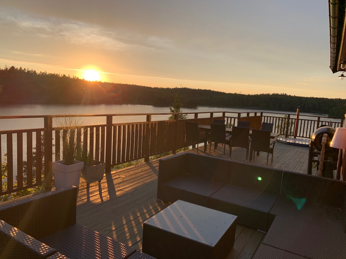 A spacious deck overlooks the tranquil waters at sunset, with silhouettes of trees in the background. Comfortable seating arrangements include woven lounge furniture. Soft light casts gentle shadows, creating a peaceful ambiance around the space.