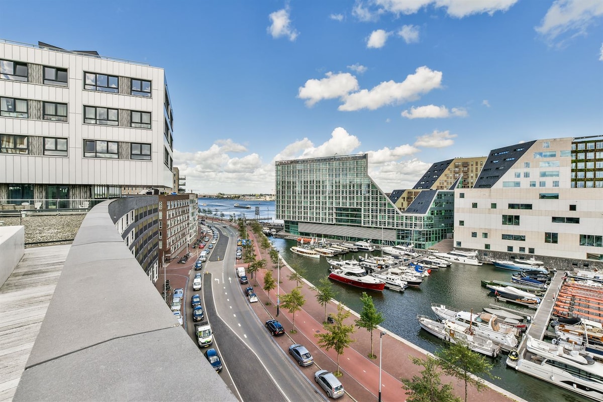 A panoramic view of a waterfront area is captured, showcasing a blend of modern buildings along a canal. Several boats are docked in the marina, while trees line the pathway beside the road. A clear sky with scattered clouds is visible above.