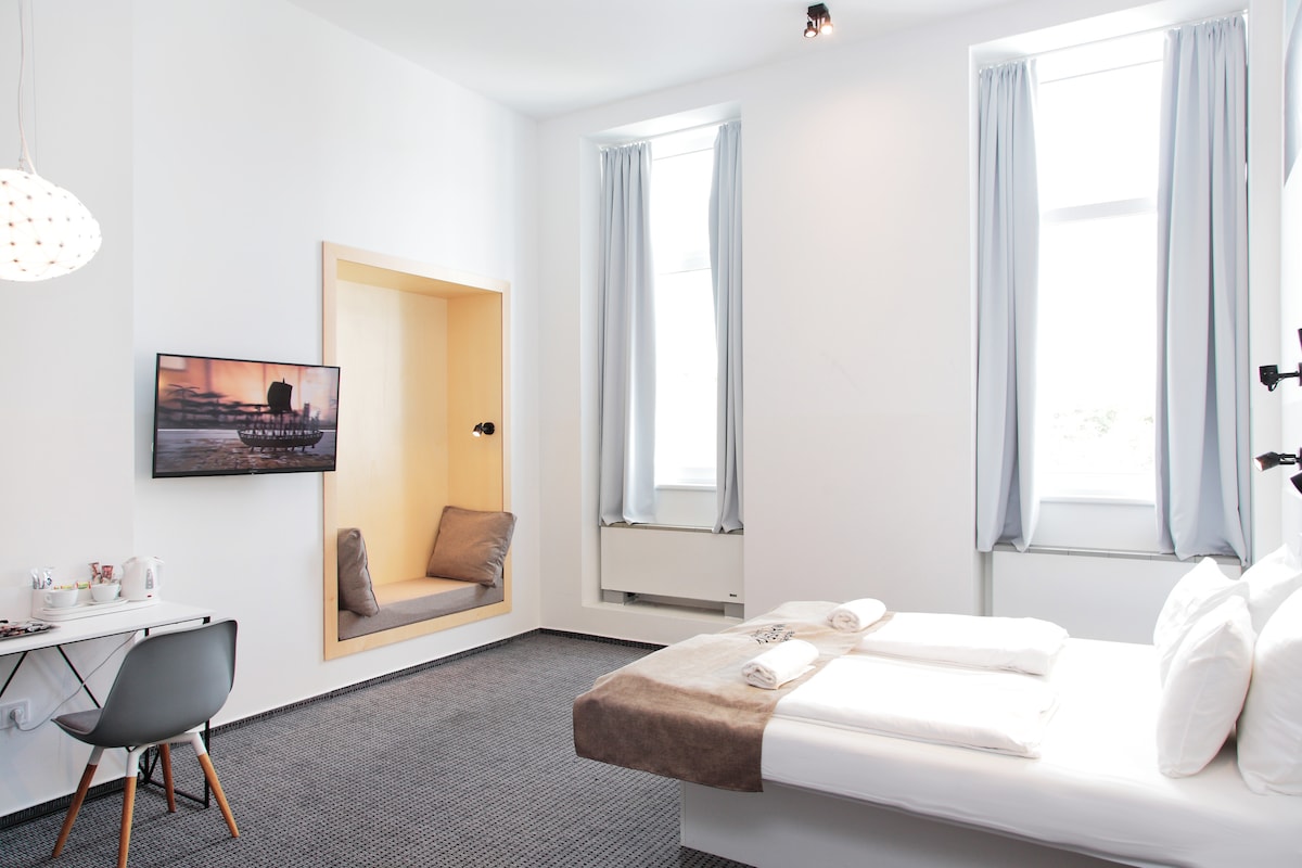 A brightly lit room features a spacious bed with neatly arranged linens and large pillows. A compact workspace with a chair is positioned under a wall-mounted television. Natural light filters through two tall windows, complemented by light drapes. A cozy nook with seating is built into the wall.