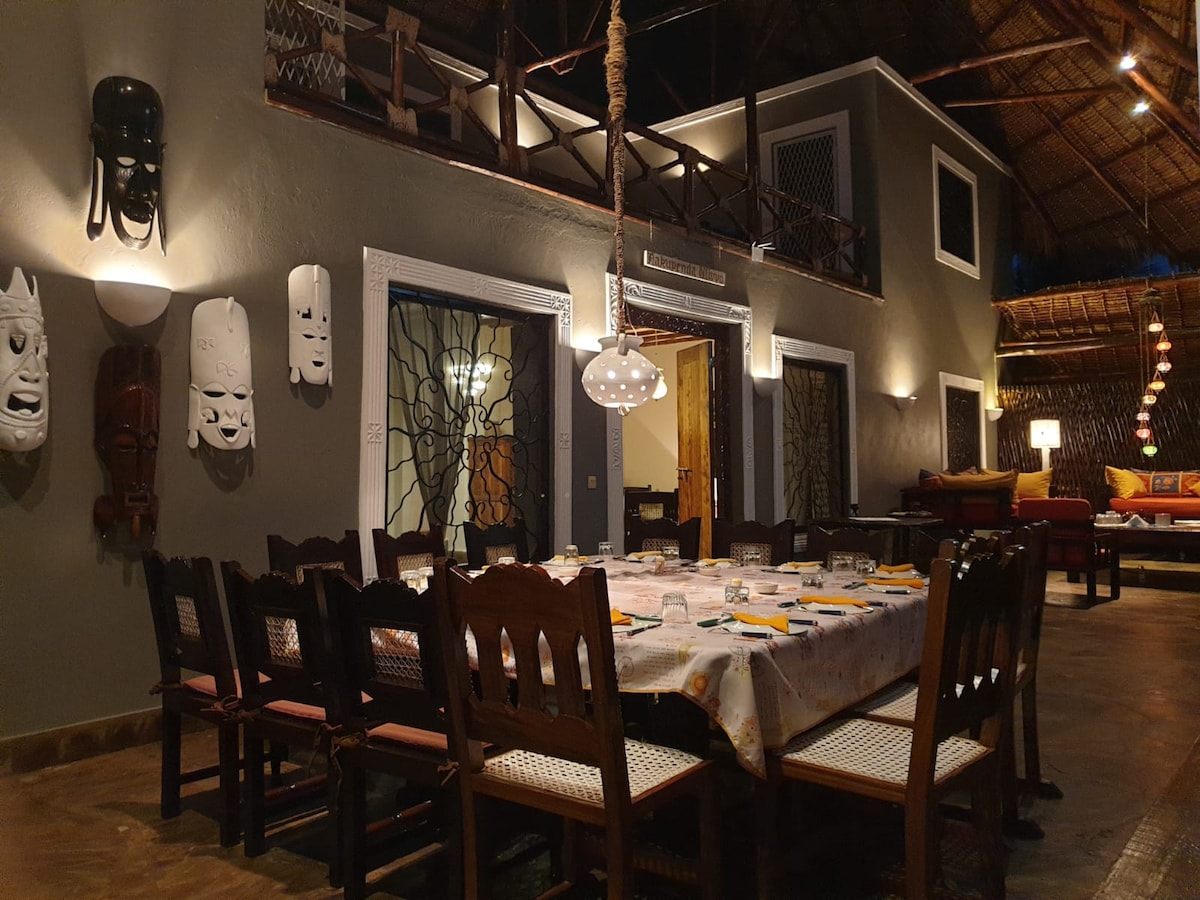 A spacious dining area is highlighted by a long wooden table surrounded by dark wooden chairs. Unique wall masks add character to the neutral-colored walls. Soft lighting enhances the ambiance, while a partition leads to a cozy seating area in the background.