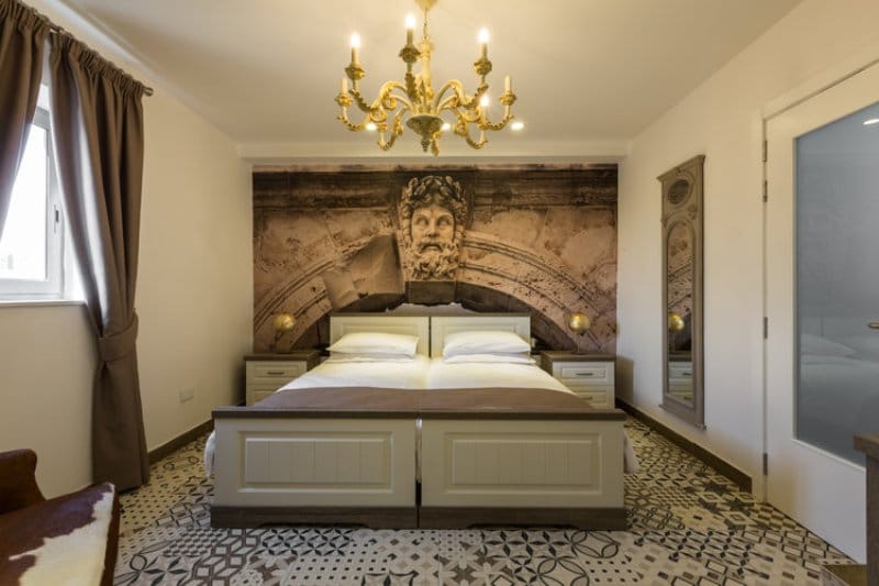A comfortable sleeping area features a spacious bed with white linens. Elegant bedside lamps provide soft illumination. A decorative chandelier hangs above, complementing the stylish wall art. The patterned floor adds character, while large windows allowed natural light to brighten the space.