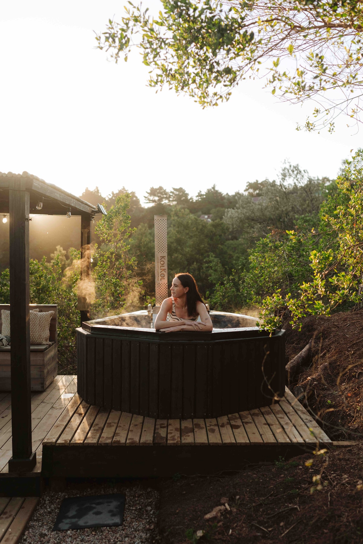 An outdoor hot tub is situated on a wooden deck, surrounded by lush greenery. Warm steam rises gently from the water. Nearby, a shaded seating area with comfortable chairs invites relaxation, while the setting sun casts a soft glow over the peaceful landscape.