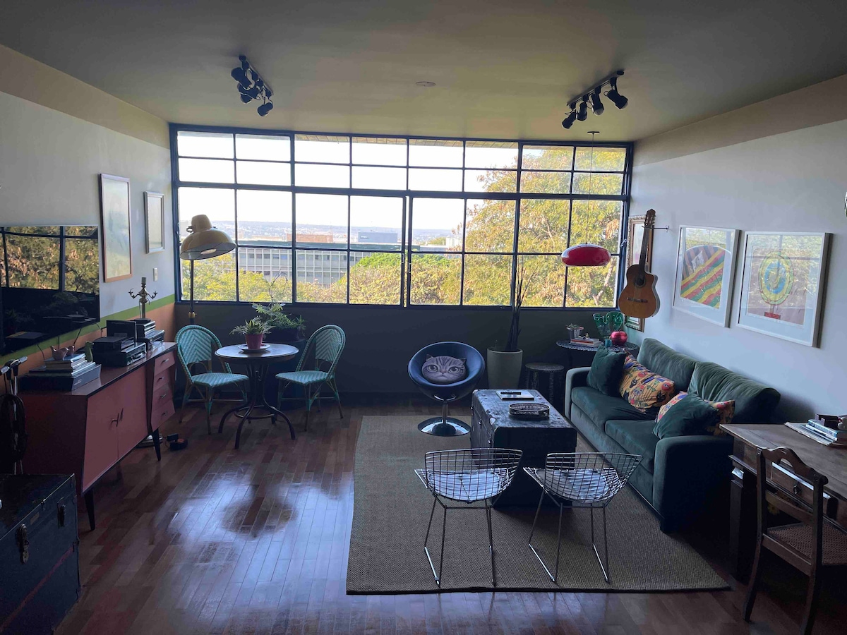 A cozy living area features large windows that provide ample natural light and scenic views. Comfortable seating includes a green sofa and accent chairs. A stylish coffee table is surrounded by artistic decor, while two dining tables invite casual meals. A guitar hangs on the wall.