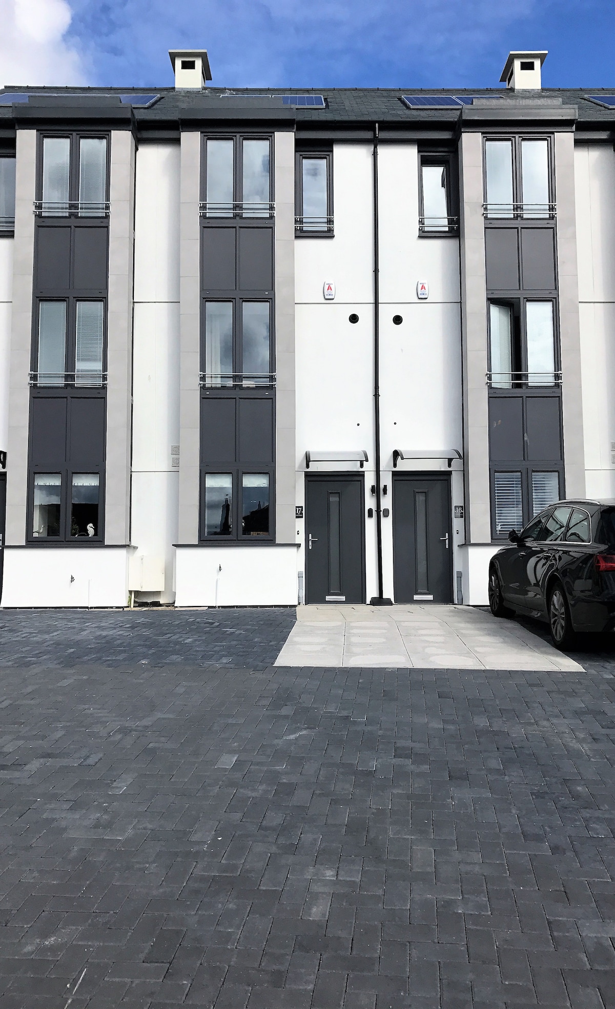 A modern, three-story building features a clean, contemporary facade with large windows and a stylish entrance. The building is flanked by a private driveway that accommodates parking for two vehicles. The grey paver stones create a neat and organized exterior space.