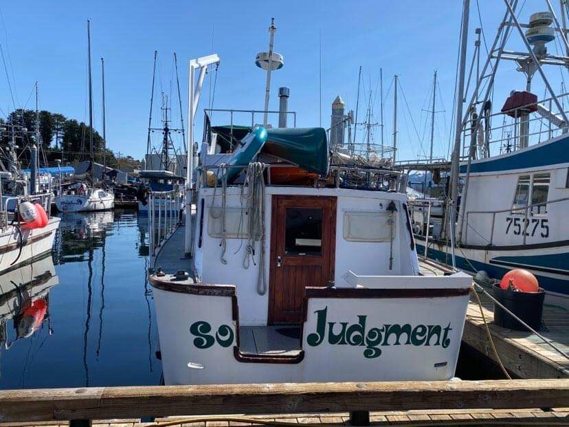The M/V Sound Judgment is docked in a bustling harbor, framed by masts of nearby boats. The boat features a well-maintained exterior with its name displayed prominently. Reflections on the water enhance the serene environment, while the clear blue sky highlights the scene.