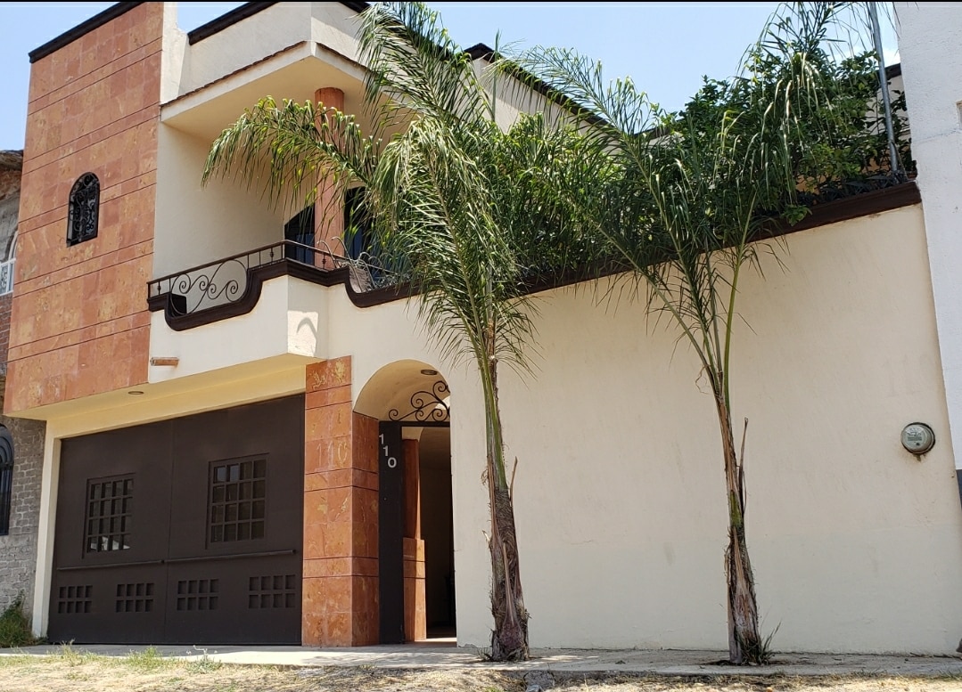 A two-story house is highlighted with a blend of warm orange and neutral stucco. Tall palm trees frame the entrance, while a large garage door indicates ample parking space. An iron railing adorns the balcony, adding a touch of charm to the exterior.