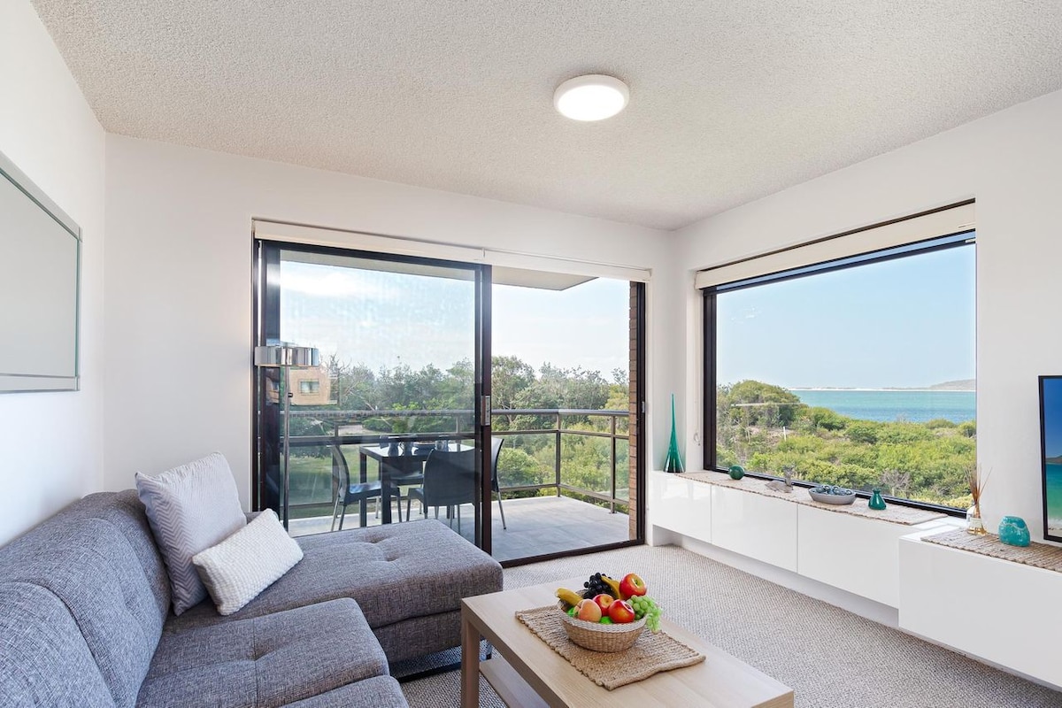 An open-plan living area features a comfortable gray sectional sofa and a light wooden coffee table adorned with a fruit bowl. Large windows provide sweeping views of the surrounding greenery and water, while a balcony with outdoor seating enhances the space.