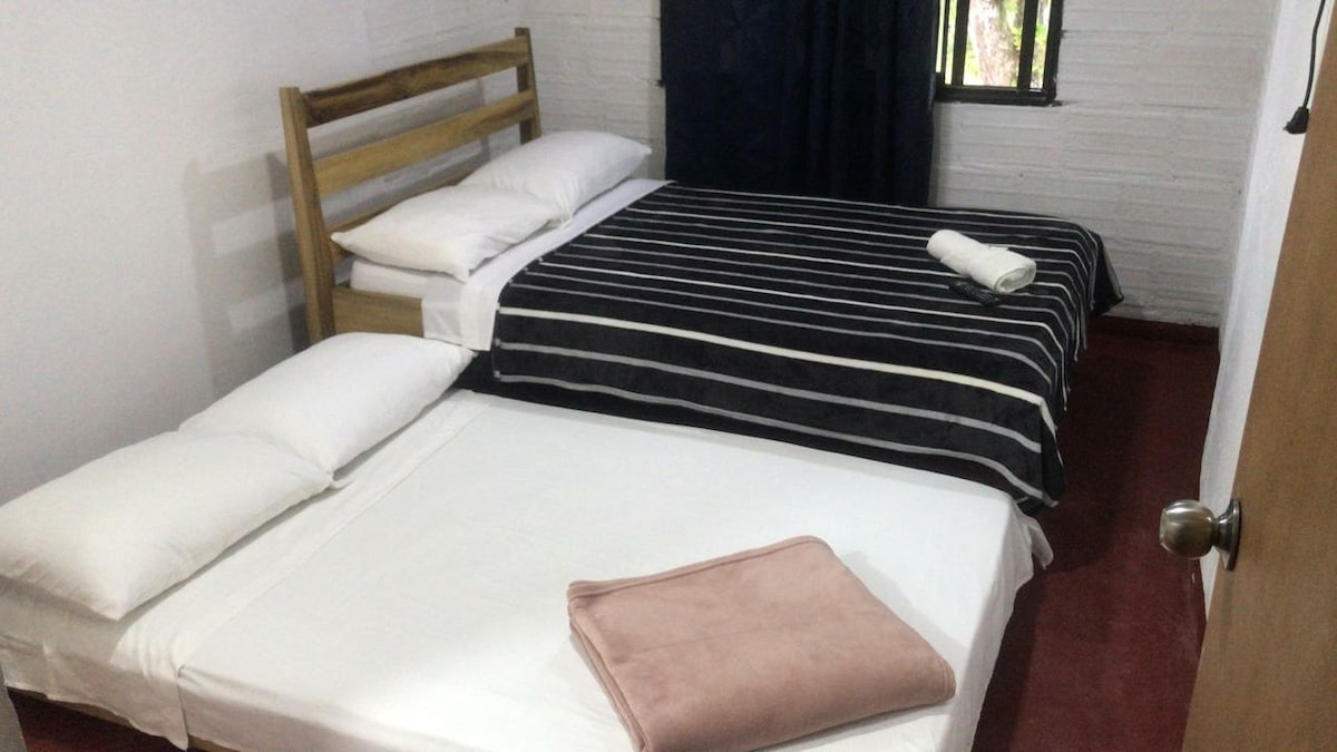 A cozy sleeping area is featured with two beds, one elevated and one positioned lower. The elevated bed has white linens, while the lower bed is dressed in a neutral blanket. Natural light filters through a window, complemented by dark curtains.