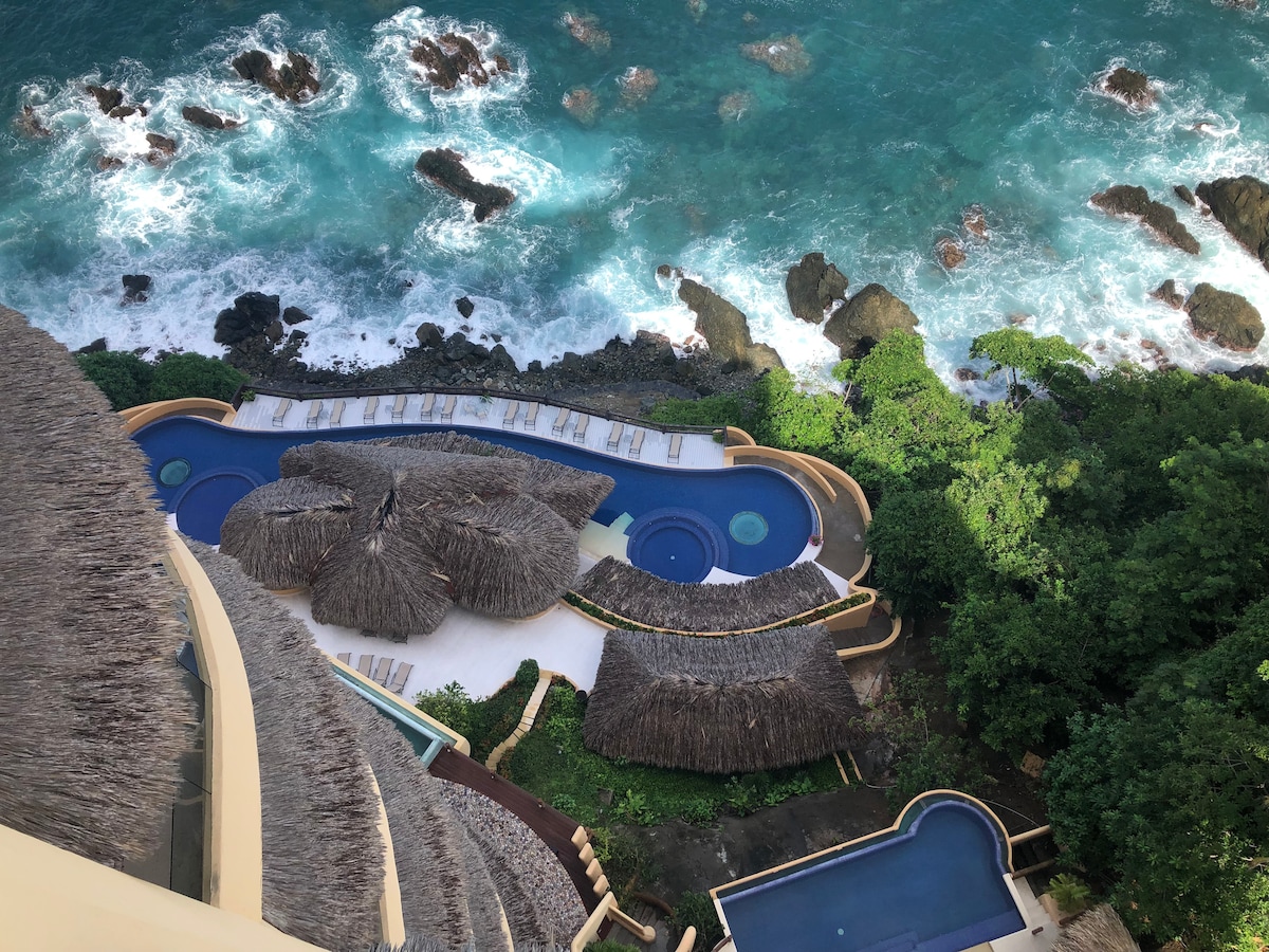 An aerial view reveals the infinity pool and jacuzzi, located near the ocean's edge. Surrounding lush greenery adds to the natural setting, while rocky shores and the vibrant blue waters of the sea create a striking contrast.