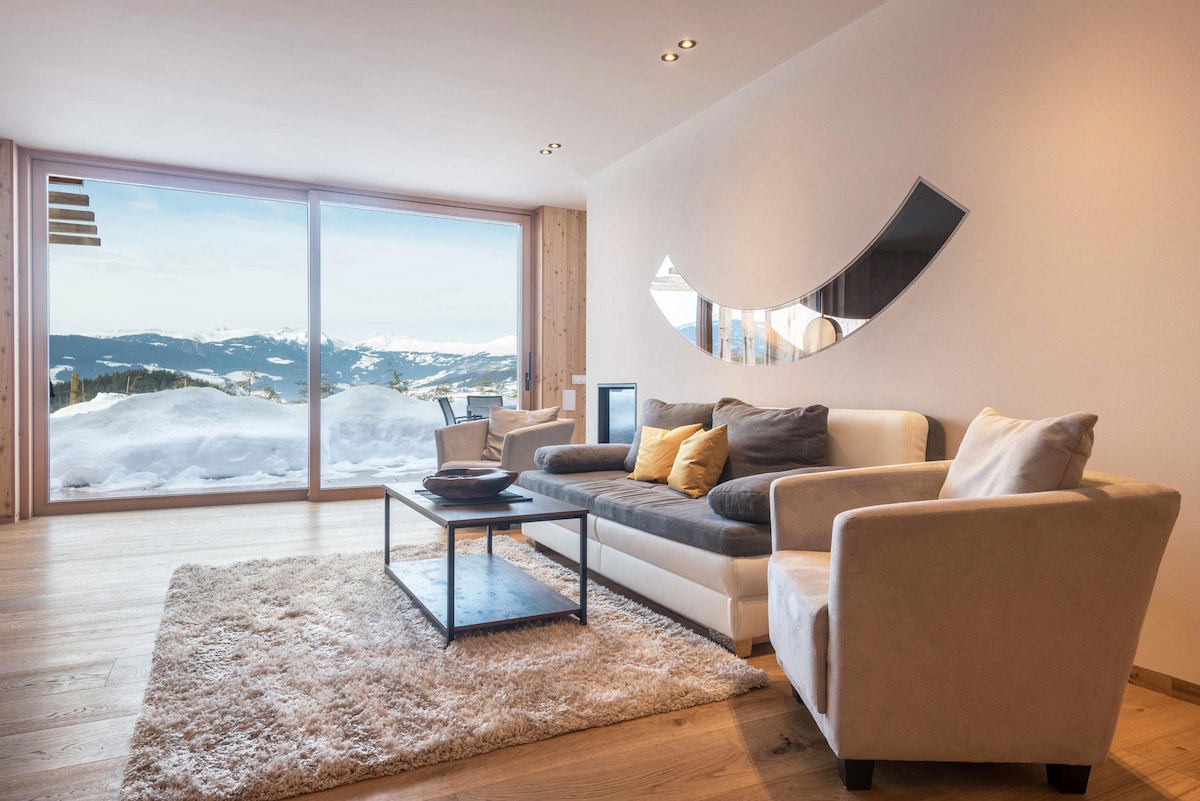 A comfortable living area features a sofa with decorative cushions, complemented by a plush area rug. A sleek coffee table sits at the center, while large glass doors provide expansive views of the snowy mountain landscape outside.
