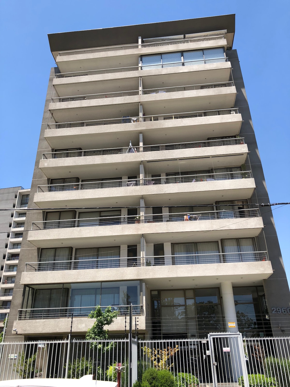 The modern building features a clean concrete façade with multiple floors and balconies. Large windows enhance natural light while a green area with shrubs is visible in the foreground. A gated entrance provides access to the property, contributing to a secure environment.