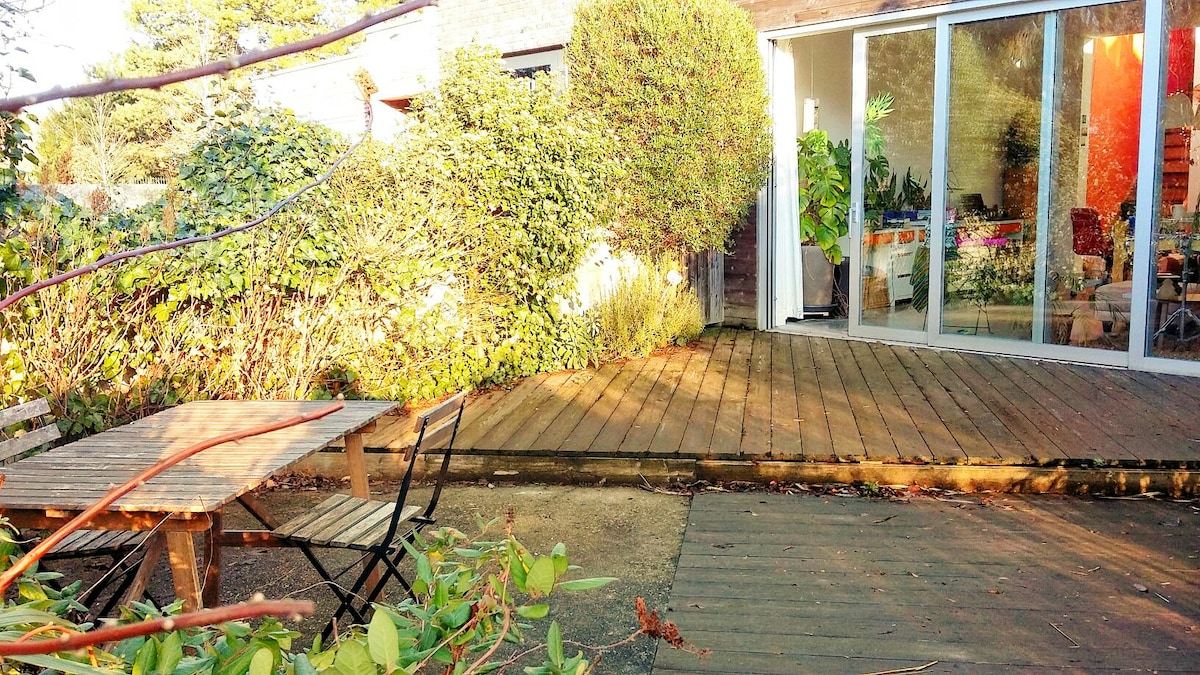 A wooden deck area is complemented by a compact outdoor table surrounded by two chairs. The terrace is bordered by lush greenery, with large sliding glass doors visible in the background, providing a view of the interior space.