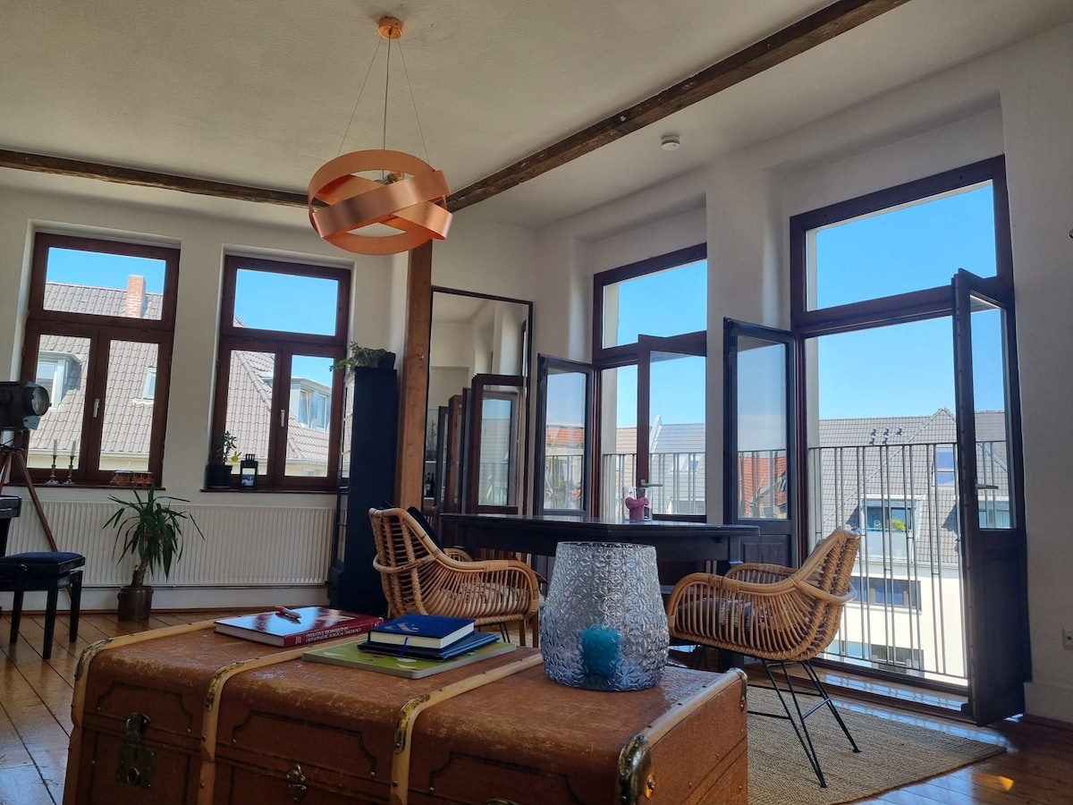 The living area features large windows that open to a bright sky, allowing natural light to fill the space. Two rattan chairs are positioned near a wooden table, complemented by decorative plants. A vintage trunk sits in the foreground, part of a cozy yet spacious environment.