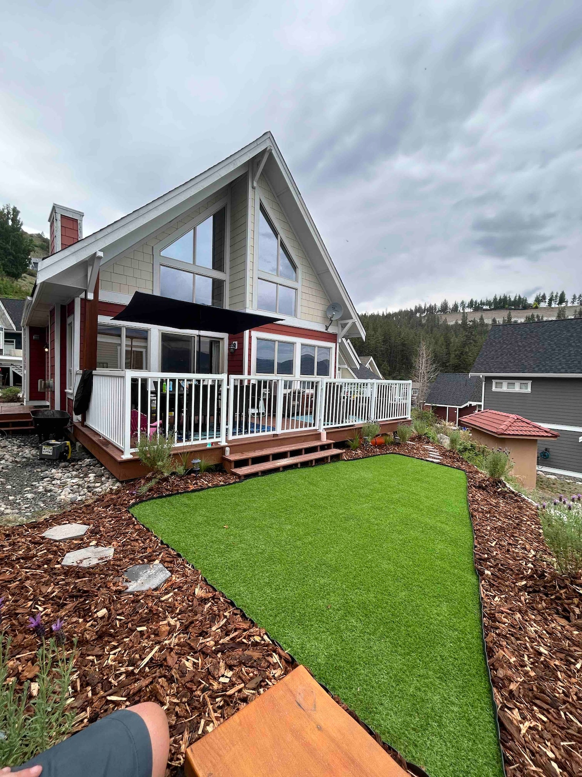 The exterior of the home features a triangular-roof design with large windows that allow for natural light. A spacious deck is visible, equipped with a shaded seating area. Surrounding the lawn, landscaped areas contain decorative stones and flower beds, enhancing the outdoor environment.