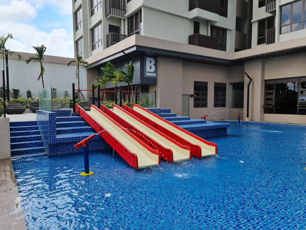 A vibrant outdoor pool area features three colorful water slides, inviting for children. The pool is surrounded by well-maintained blue tiles and is accented by tropical plants. Shallow steps lead into the water, enhancing accessibility and enjoyment.
