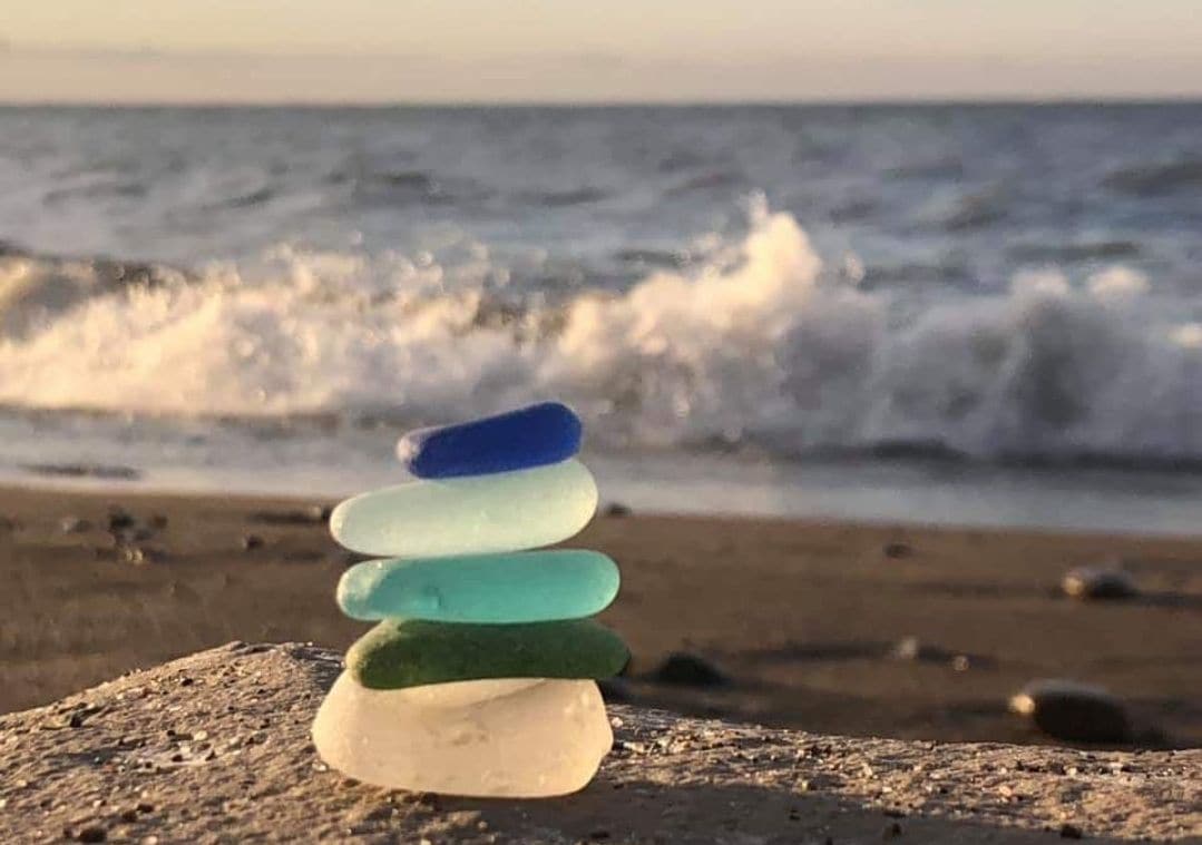 A stack of sea glass in varying shades of blue and green is positioned on a sandy shore. The background features gentle waves lapping against the beach, with a soft horizon reaching towards the ocean under a pale sky.