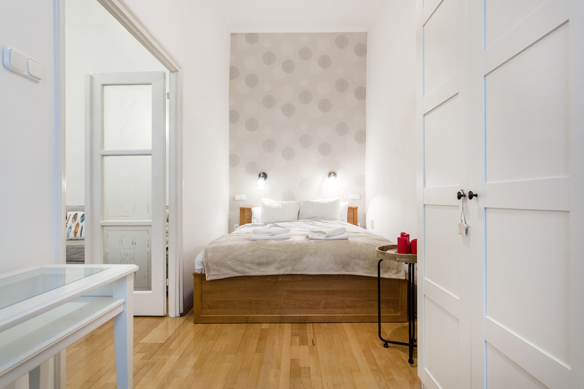 A serene bedroom is visible, featuring a spacious bed with neatly arranged white linens. Soft lighting is provided by two wall-mounted lamps on either side. A small side table holds decorative candles, adding a subtle touch to the warm wooden flooring.