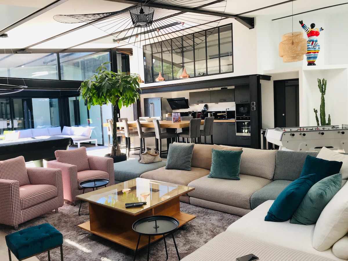 A spacious living area features a large sectional sofa with a combination of neutral and teal cushions. A center coffee table sits atop a soft area rug, while a dining area with high chairs is visible in the background. Large windows provide ample natural light.