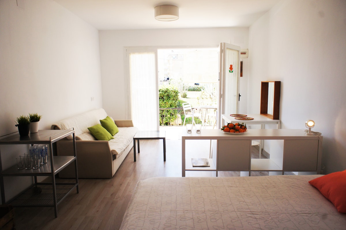 The interior features a spacious bed and a light-filled seating area with a sofa adorned with cushions. A dining table is positioned near glass doors leading to a private terrace, which is furnished with chairs. Fresh fruit is displayed on the table.