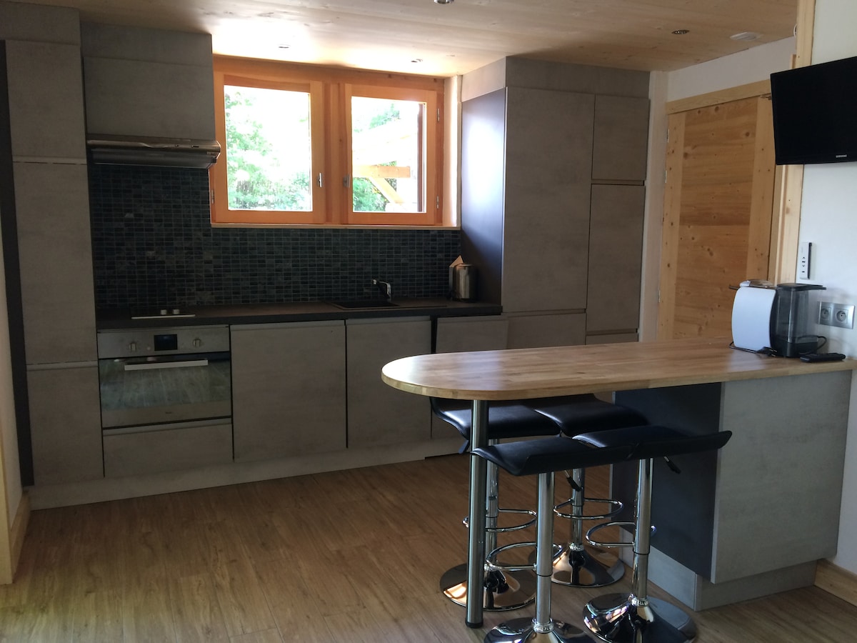 A modern kitchen is equipped with essential appliances and a sleek countertop. Natural light enters through large windows, creating a bright environment. The cabinetry showcases a minimalist design, complemented by a small dining area with barstools. Wood accents add warmth to the space.