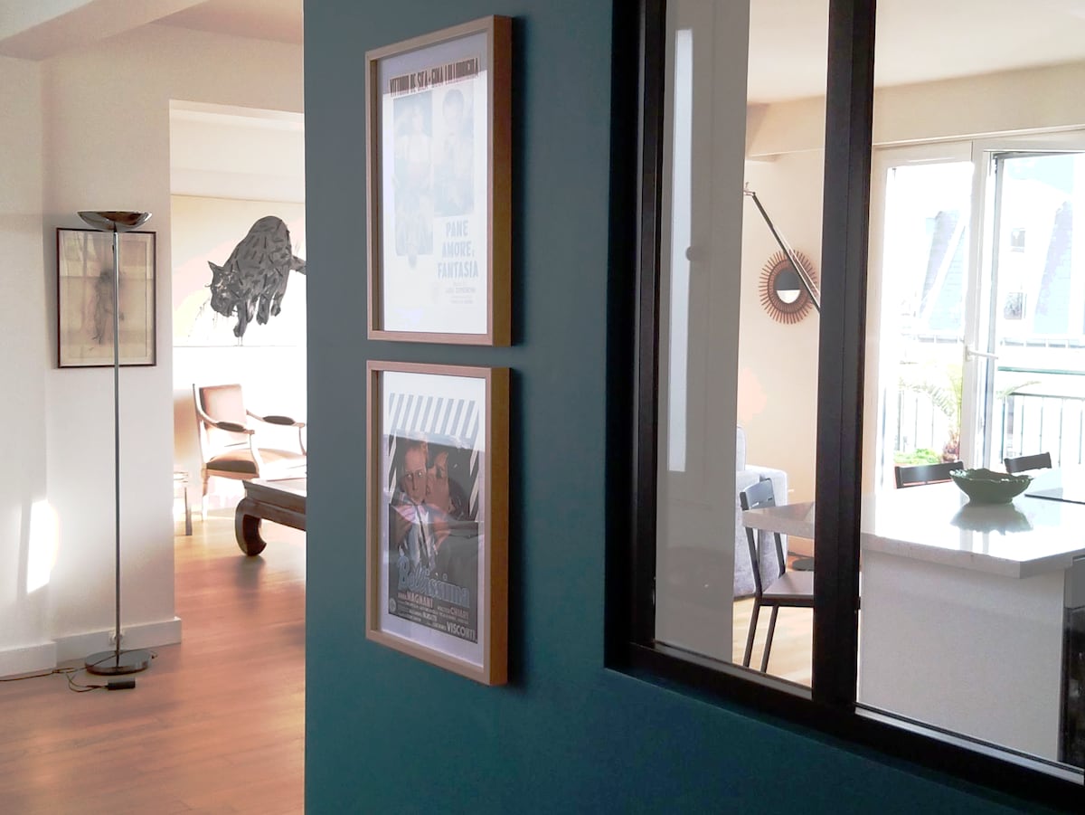 A corridor painted in a soothing teal color leads to a bright living area. Framed artwork adorns the wall, adding a touch of character. Natural light filters through glass doors, highlighting the warm wood flooring and inviting atmosphere.