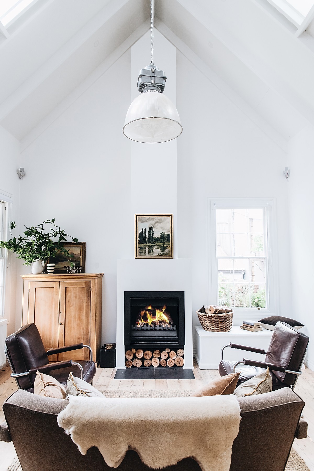 A bright lounge area features a high, sloped ceiling and warm natural light. A central fireplace, surrounded by neatly stacked logs, provides a focal point. Two leather armchairs and a wooden cabinet are complemented by a cozy throw blanket and decorative items on the shelves.