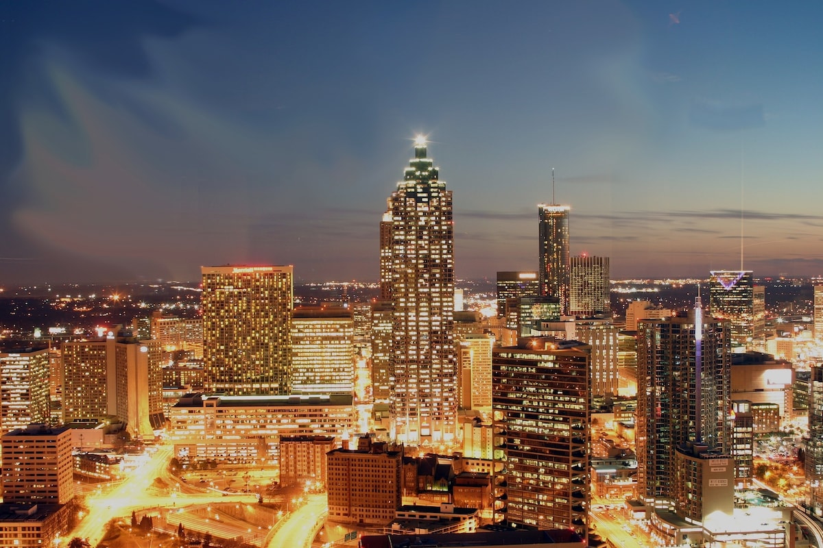 A vibrant city skyline is captured during dusk, with various buildings lit up in a mix of colors. The tallest structure stands prominently, its peak illuminated. The horizon is dotted with lights, creating a dynamic urban atmosphere against a fading blue sky.
