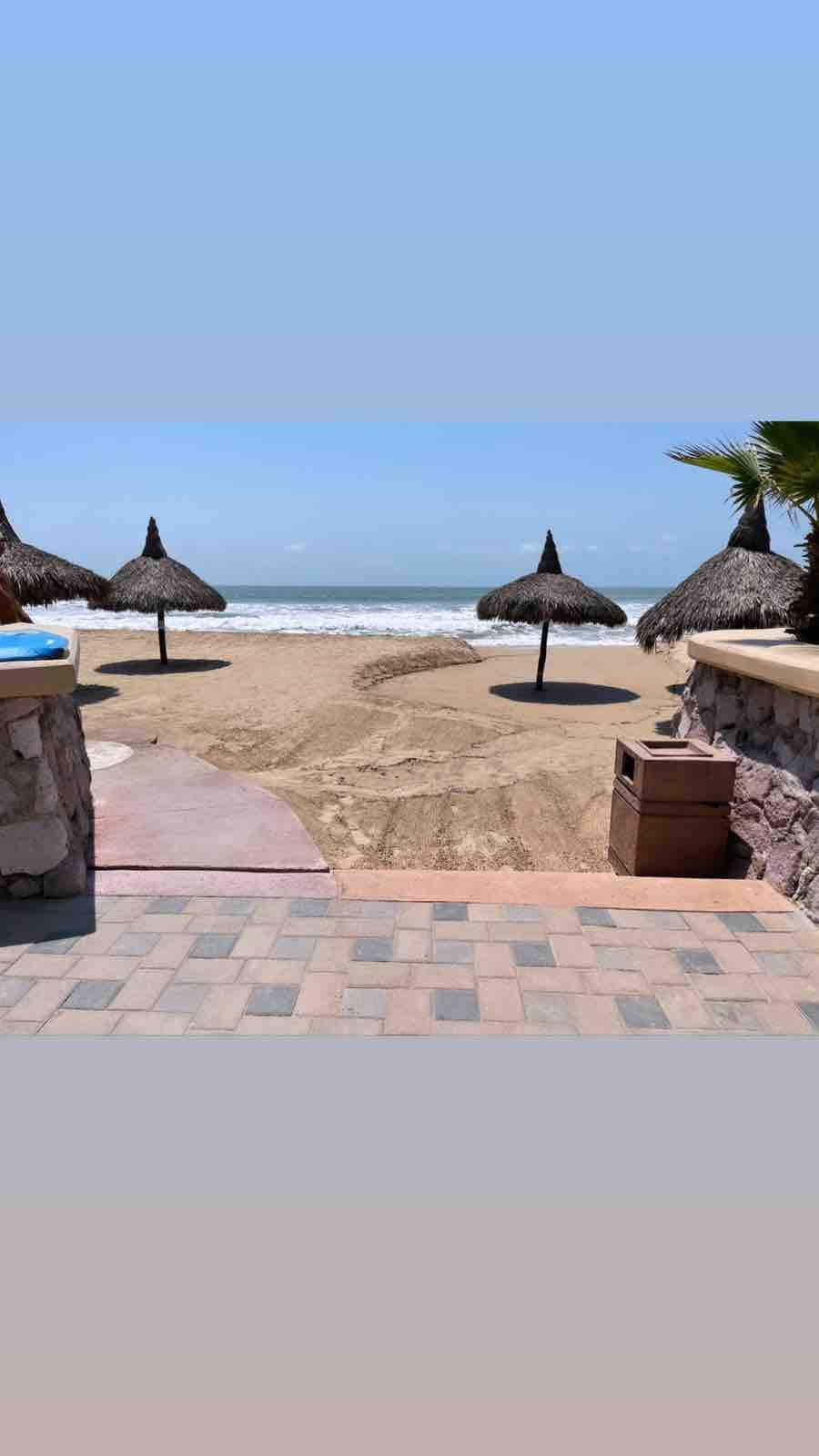 The image presents a serene beach scene with soft sand and gentle waves. Palapas with thatched roofs provide shaded areas on the beach, while a stone pathway leads from the foreground to the shoreline, offering direct access to the ocean.