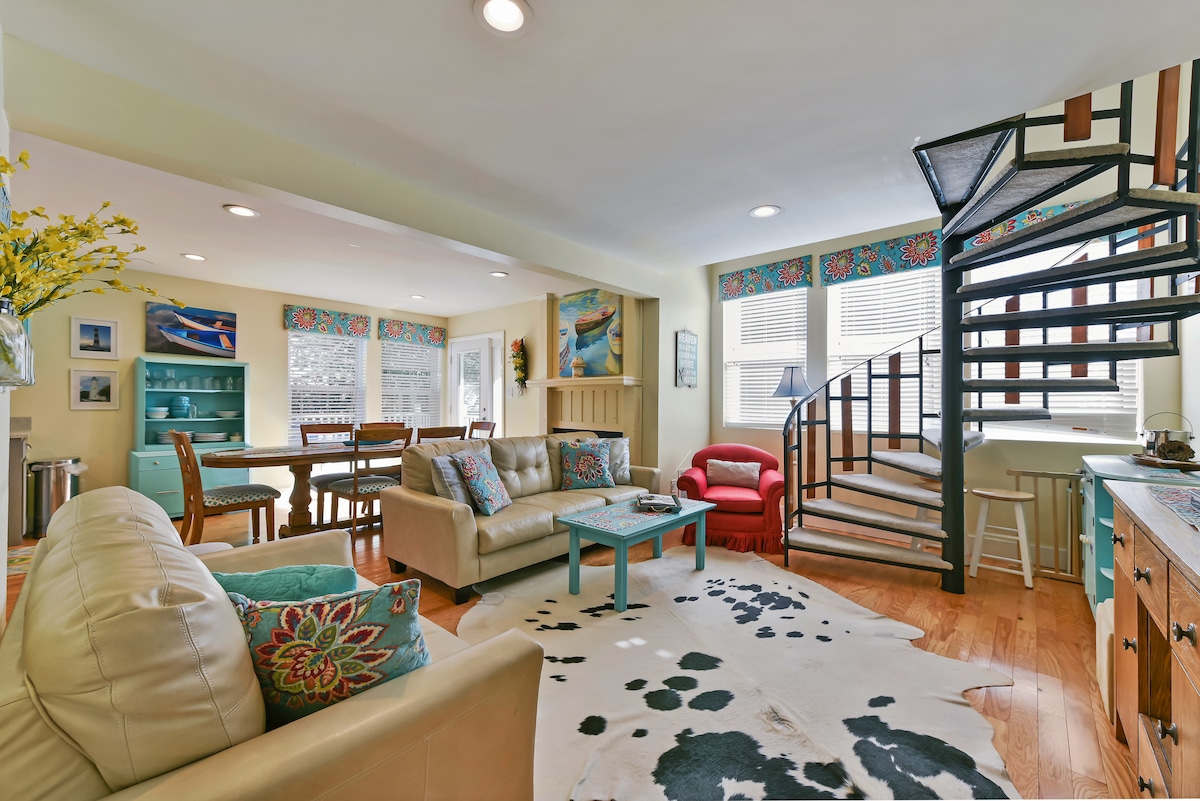 An open living area features light-colored leather seating, adorned with decorative pillows. A coffee table sits centrally, paired with a patterned rug. A dining table accommodates several chairs, while a spiral staircase provides access to the upper level. Natural light filters through covered windows.
