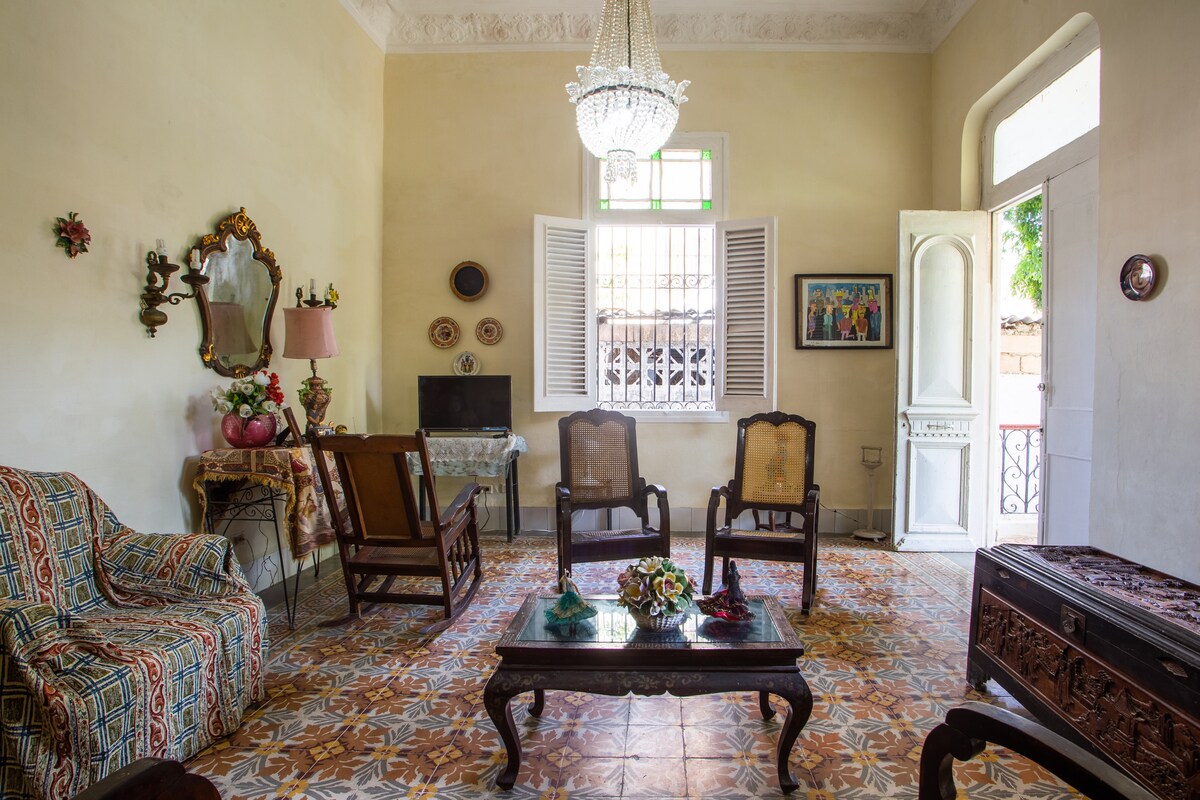A spacious living area is highlighted by traditional furniture, including two armchairs and a patterned sofa. A coffee table is centered on a tiled floor, while a small television and decorative items are visible on a side table. Natural light filters through the windows, enhancing the room's ambiance.
