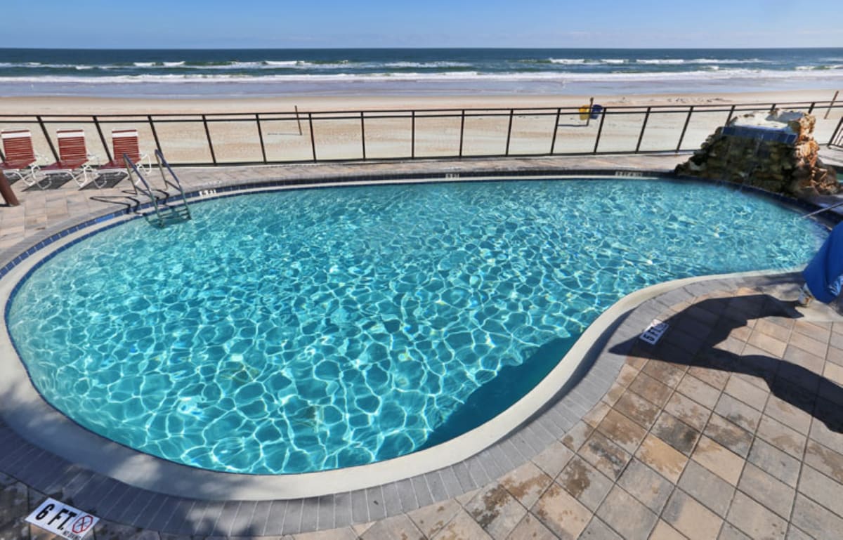 A curved pool is set against a backdrop of the ocean, featuring clear blue water reflecting sunlight. Beach chairs arranged near the poolside provide seating options, with the sandy shore and waves visible in the distance.