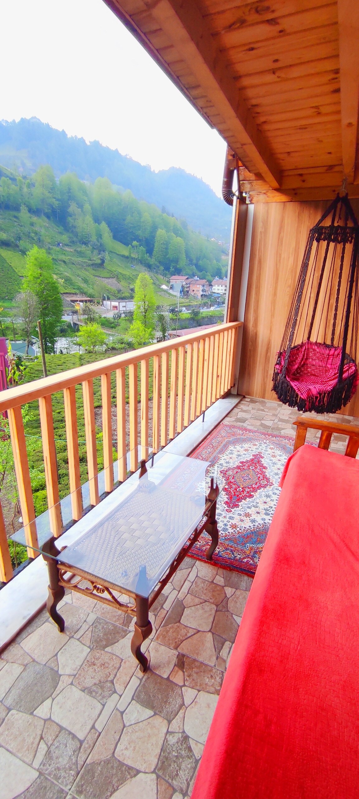 A balcony is shown with wooden railings offering a view of lush green hills. A glass table rests on a stone floor, complemented by a colorful area rug. A hanging chair adds a cozy seating option, inviting relaxation in a serene outdoor setting.
