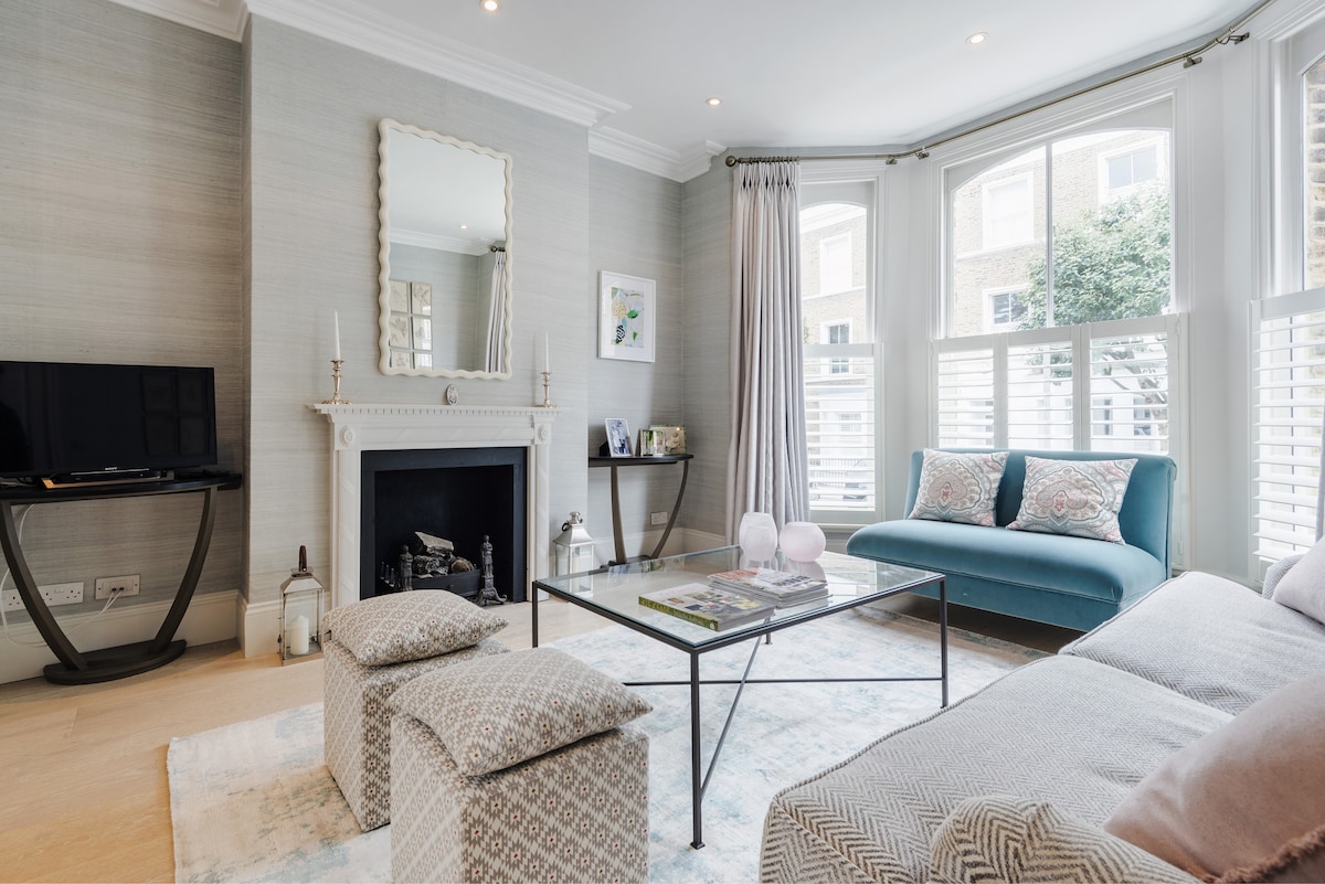A well-appointed living area features a light-colored sofa and a glass coffee table surrounded by patterned poufs. A wall-mounted television is positioned beside a decorative fireplace, and large windows with shutters offer natural light and views of greenery outside.