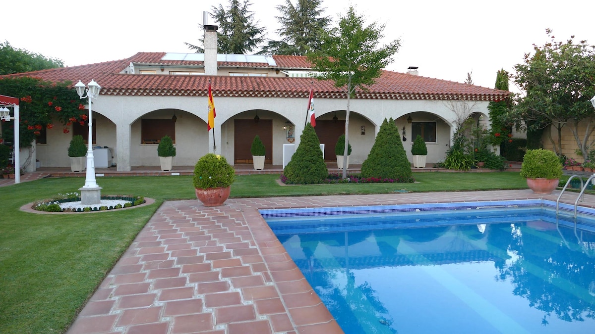 The villa's exterior features a well-maintained garden with manicured hedges and vibrant flower beds. A spacious swimming pool glistens in the foreground, while a charming tiled walkway leads to the entrance, framed by archways and potted plants. The building displays a traditional roof and decorative flags.