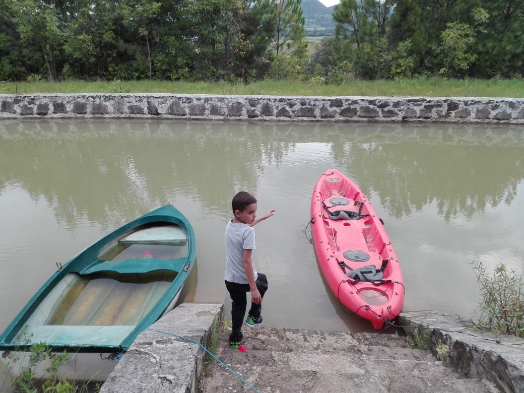 A child stands on stone steps leading to a calm, reflective body of water. Two kayaks are anchored at the water's edge, with one in vibrant red and the other in a muted green. Verdant trees are visible in the background, adding to the serene setting.