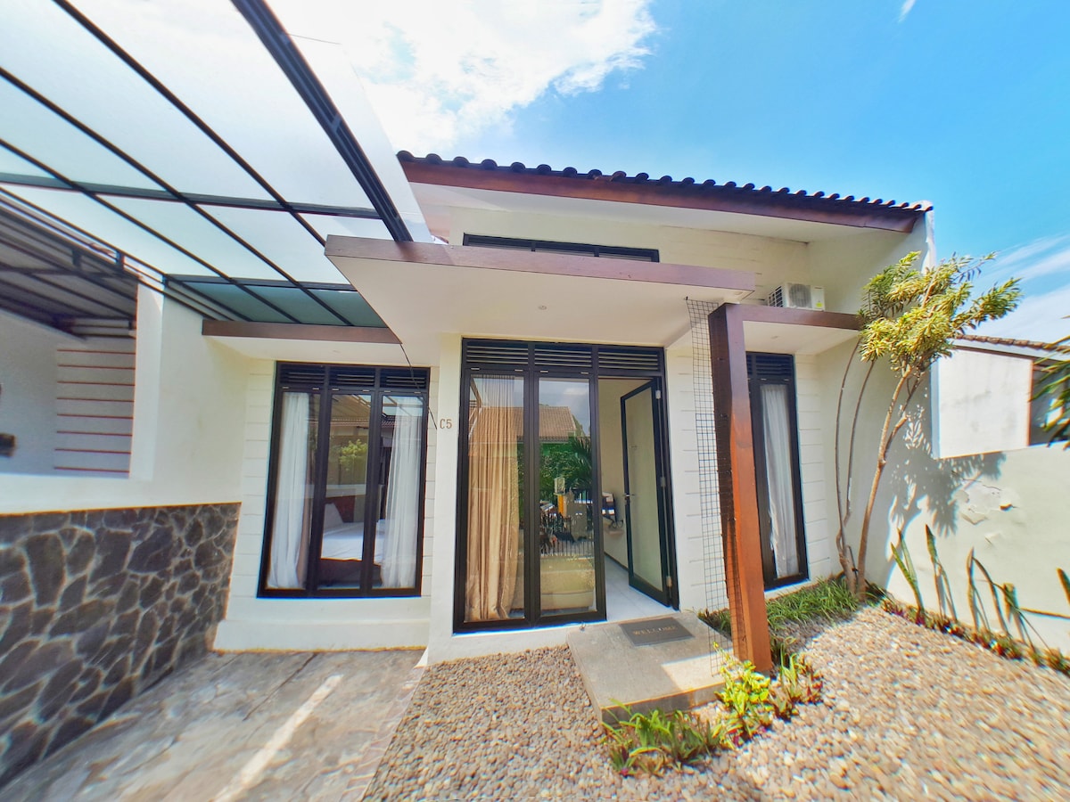 The villa's exterior showcases a modern minimalist design with large glass doors that allow natural light to enter. A stone pathway leads to the entrance, surrounded by neatly arranged gravel and greenery. The roof features a transparent overhang, blending interior and exterior spaces.