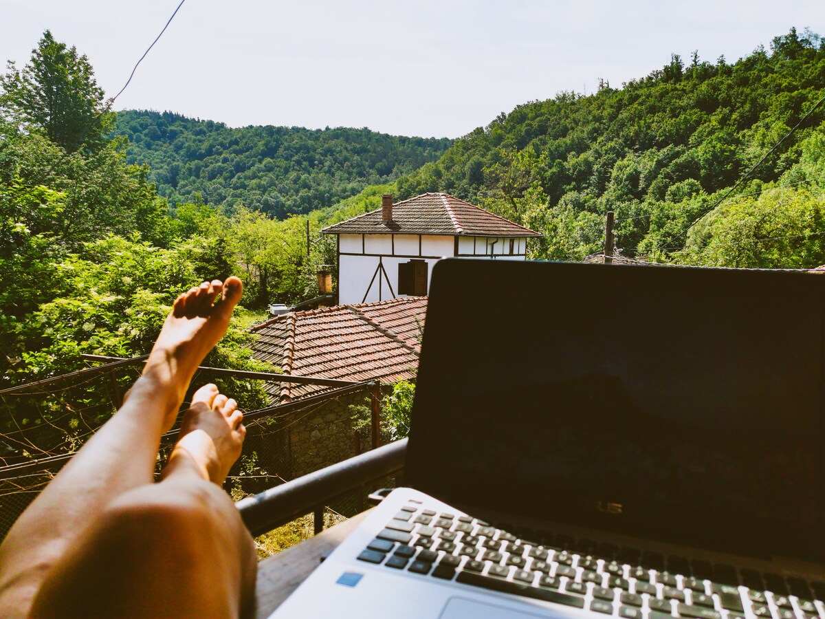 A scenic view from a balcony showcases lush green mountains and a historic building in the distance. Relaxed feet can be seen resting on the railing, while a laptop sits open on a table, suggesting a blend of work and leisure in nature.