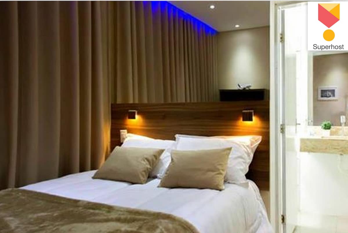 The sleeping area features a spacious bed with soft linens and neutral-toned pillows. Warm lighting illuminates the space, accentuated by a backdrop of smooth, draped fabric. A subtle design element is visible in the adjoining area, suggesting a modern aesthetic.