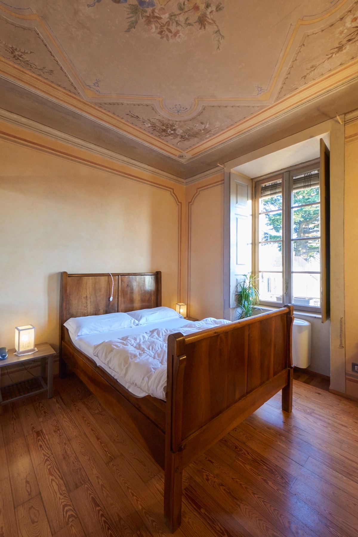 A warm-toned room features a spacious wooden bed with a simple white bedding arrangement. Original ceiling frescoes highlight the historic charm. Natural light enters through the windows, illuminating the wooden floor and antique nightstands beside the bed.