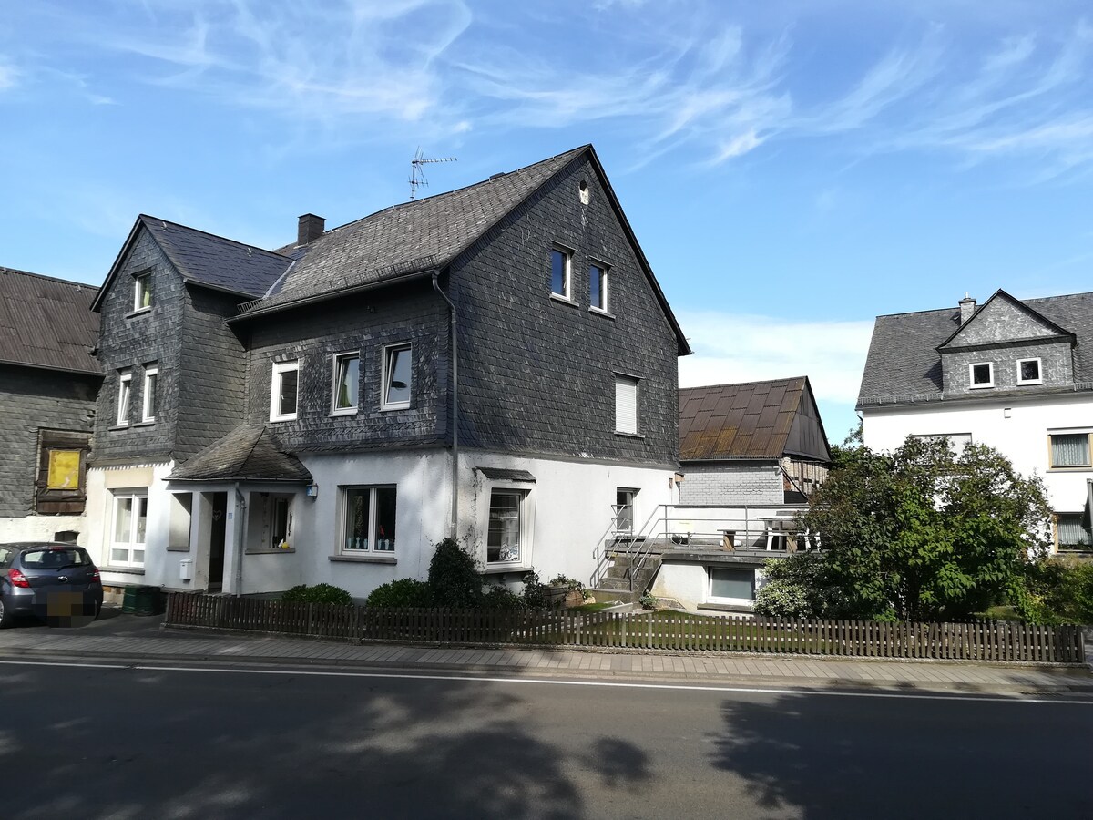 A two-story house is seen from the street, featuring a combination of dark stone and white walls. Windows are adorned with flower boxes, and a small front yard is enclosed by a wooden fence. A patio area is visible on the side of the building.