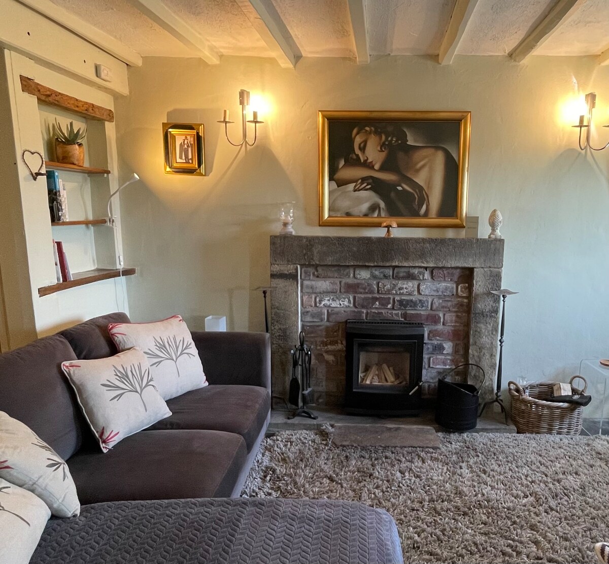 A cozy lounge area features a comfortable couch adorned with decorative cushions. A stone fireplace holds a log burner, framed by a captivating artwork. Soft lighting from wall sconces enhances the inviting atmosphere. A plush rug adds warmth to the stone floor.