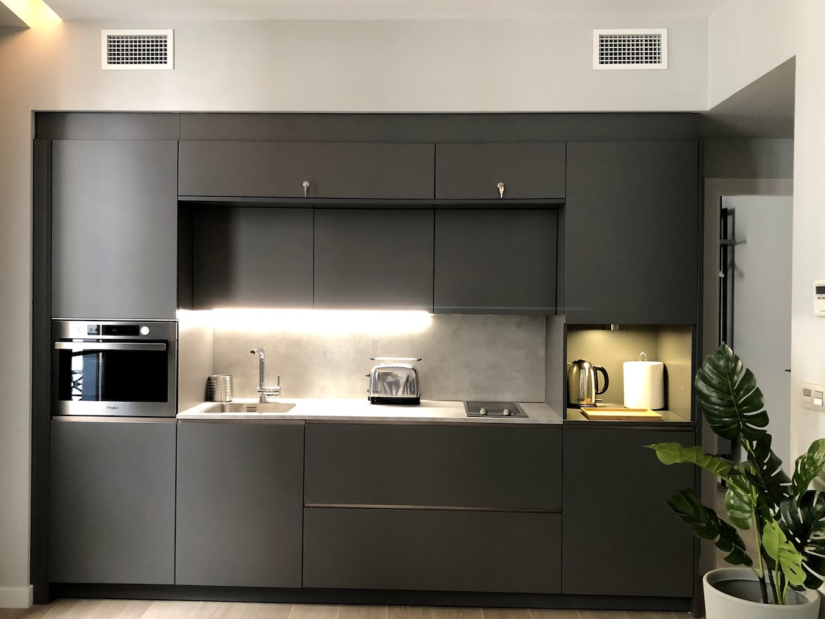 A modern kitchen space features sleek gray cabinetry, equipped with a built-in oven, cooktop, and sink. Under-cabinet lighting illuminates the countertop, which includes a toaster and kettle. A small potted plant adds a touch of greenery to the environment.