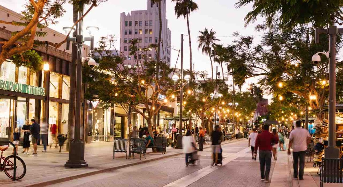 A vibrant outdoor promenade is lined with palm trees and warm street lights. People can be seen strolling along the pathways, with shops and cafes on either side, creating a lively atmosphere in a welcoming urban environment.