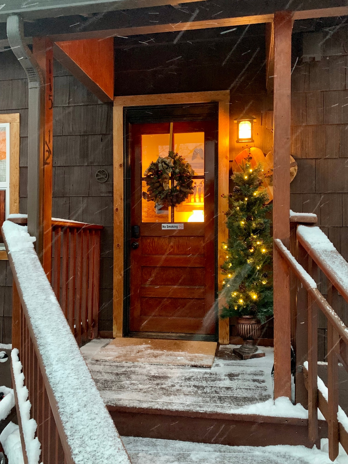 The entryway features a warmly lit door surrounded by snow-covered steps. A decorated evergreen tree stands to the right, and a wreath adorns the door, creating a welcoming appearance amidst a gentle snowfall.